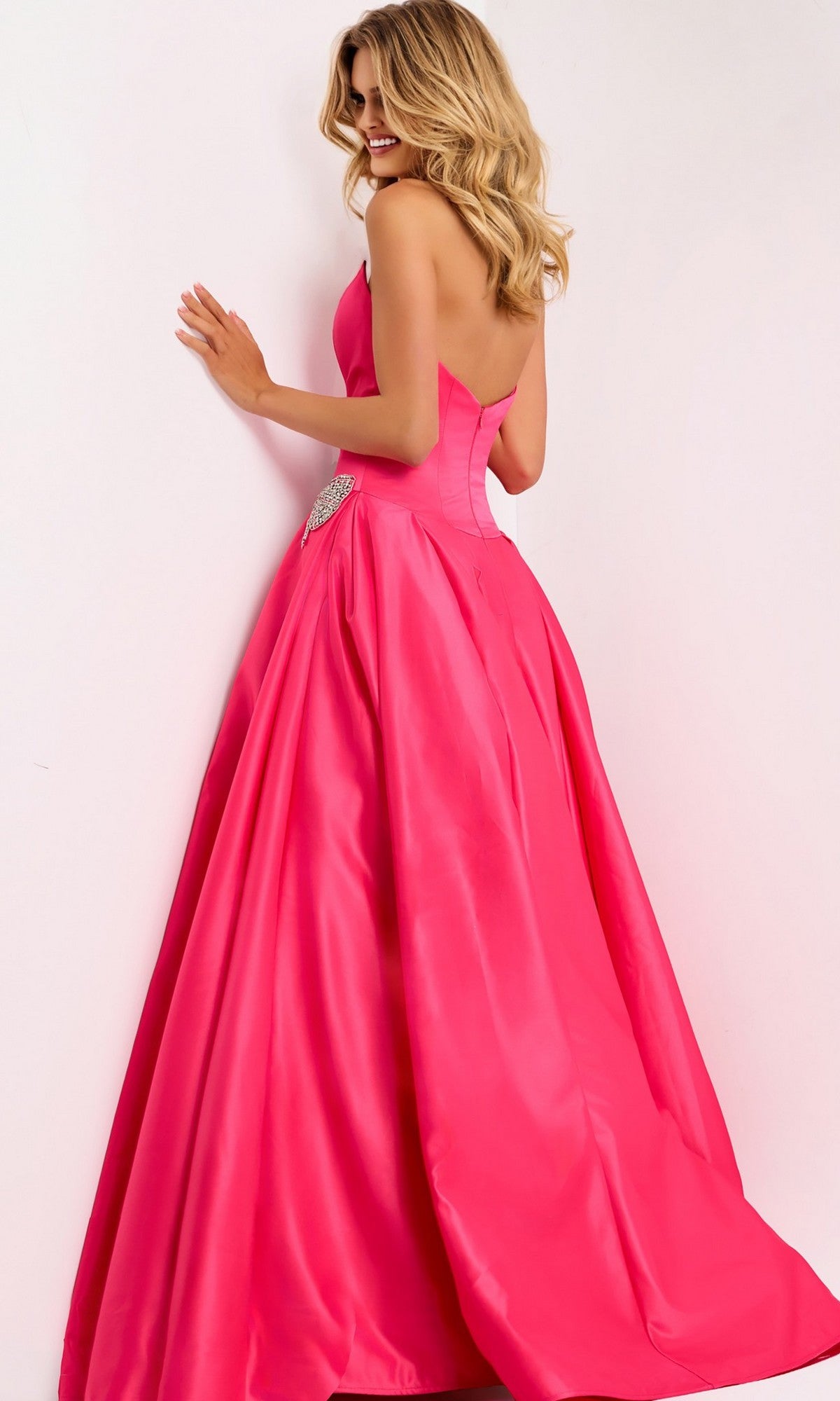A woman with wavy blonde hair models the Jovani 47096, a strapless satin long prom dress with a fitted bodice, flared skirt, and rhinestone bow, standing against a plain light background.